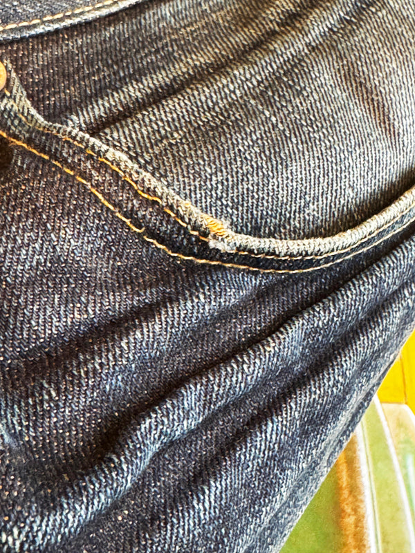 Close-up of dark indigo denim fabric showing the texture of the twill weave and a yellow-orange contrast stitch along the pocket edge.