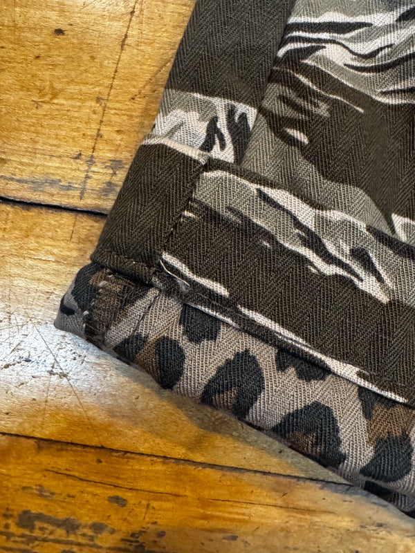 Close-up of a folded textile item showing two fabric patterns: a dark brown and gray herringbone camo section and a matching leopard-print section, laid on a rustic wooden surface.