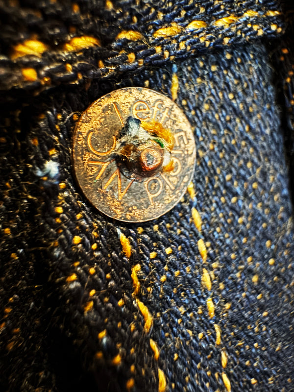 Close-up of a heavily worn, rusted button on dark blue denim with bright yellow contrast stitching.