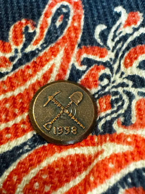 A round bronze-colored metal button featuring crossed shovel and pickaxe symbols and the year '1998', shown on a patterned fabric background with red, navy, and cream stripes.
