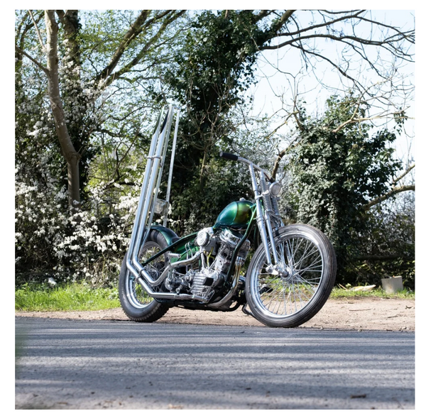 A custom chopper motorcycle with a green metallic fuel tank, chrome engine components, and a tall rear rack, parked on an asphalt road with trees in the background.
