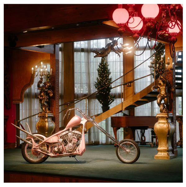 A custom chopper motorcycle with a glossy pink body and chrome details, parked on a dark green carpeted stage inside a grand, ornate hall with a staircase, chandeliers, and decorative statues.