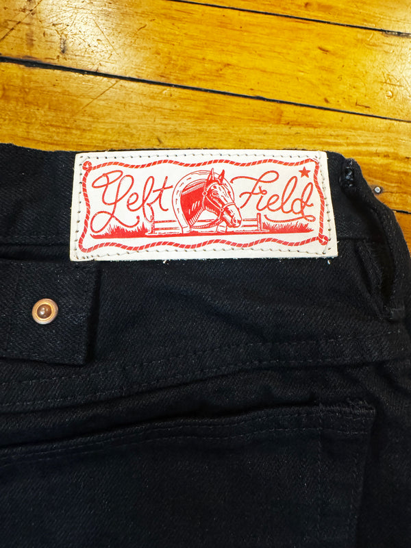 Close-up of the waistband of black denim jeans with a white and red 'Left Field' brand label, placed on a yellow wooden floor.