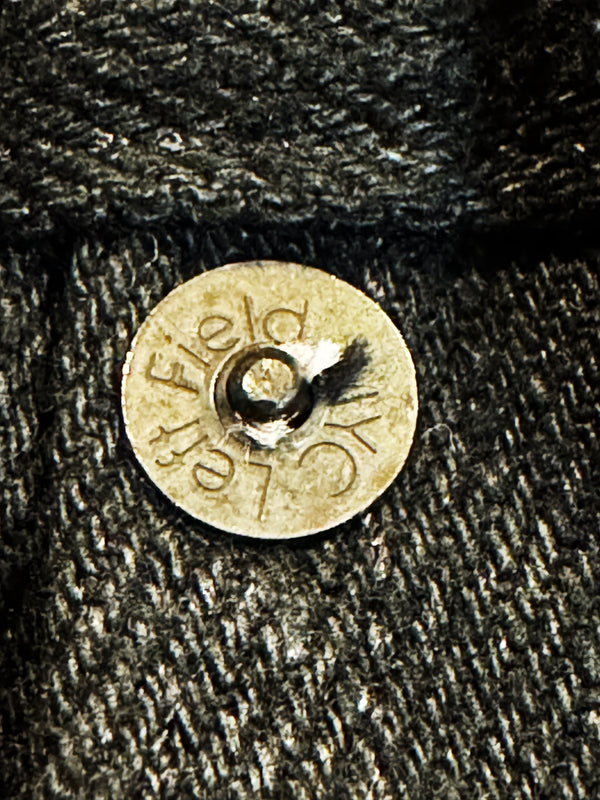 A close-up of a round brass button stamped with the words 'Left Field' on a dark textured denim fabric.