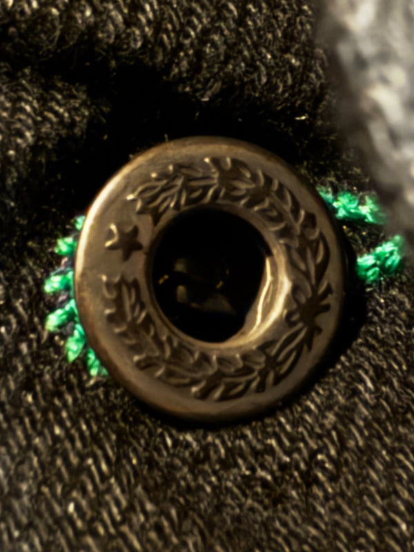 Close-up of a round bronze-toned metal button with engraved leaf and star motifs, set into dark textured fabric with bright green thread accents.