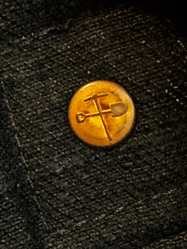 A round gold-toned metal button with an engraved crossed shovel and pickaxe symbol, set onto a dark textured fabric background.