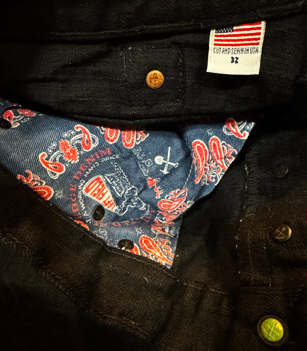 Close-up of the interior waistband of a pair of black denim jeans, showing a white size tag that reads 'CUT AND SEWN IN USA 32' and a blue denim lining with a red and white paisley pattern.