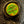 A close-up of a round vintage button with a yellow-gold background and a green patina, featuring a raised crossed pickaxe and shovel symbol. The button is set in a dark circular frame and mounted on a dark, textured fabric.