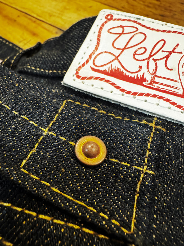 Close-up of dark indigo denim fabric with yellow contrast stitching, a wooden button, and a white sewn label printed with red script reading "Left".