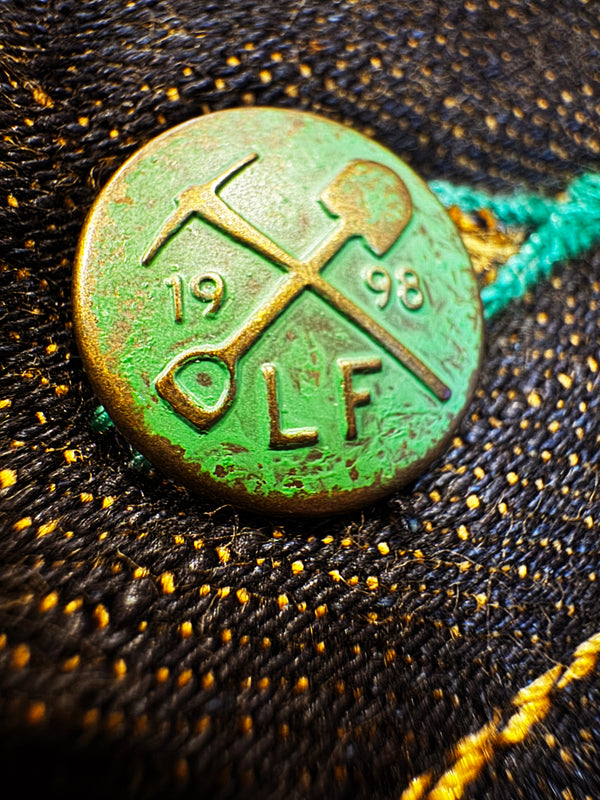 A close-up of a round vintage-style metal button with a distressed green patina and gold-toned details, featuring crossed tools and the letters 'LF' with '1998' on a black denim fabric background.
