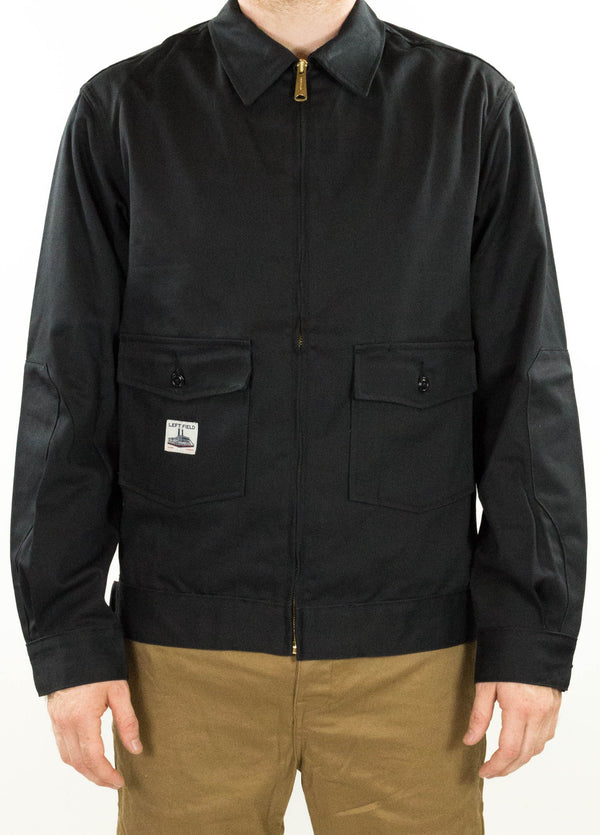 A person wearing a black twill garage jacket with a brass zipper and reinforced elbow patches.